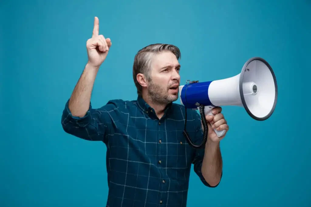 middle age man with grey hair in dark color shirt shouting in megaphone pointing with index finger up standing over blue background